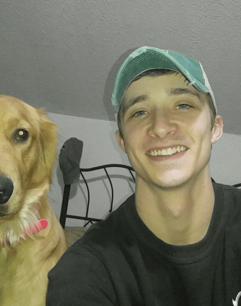 Kevin Hartley wears a green baseball hat and black shirt as he smiles. His dog Chevelle, which looks like a golden retriever sits next to him. 