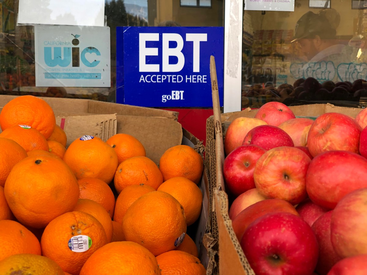 A box of oranges and a box of apples are displayed outside of a grocery store. On the window is a sign that says the store accepts EBT and WIC.
