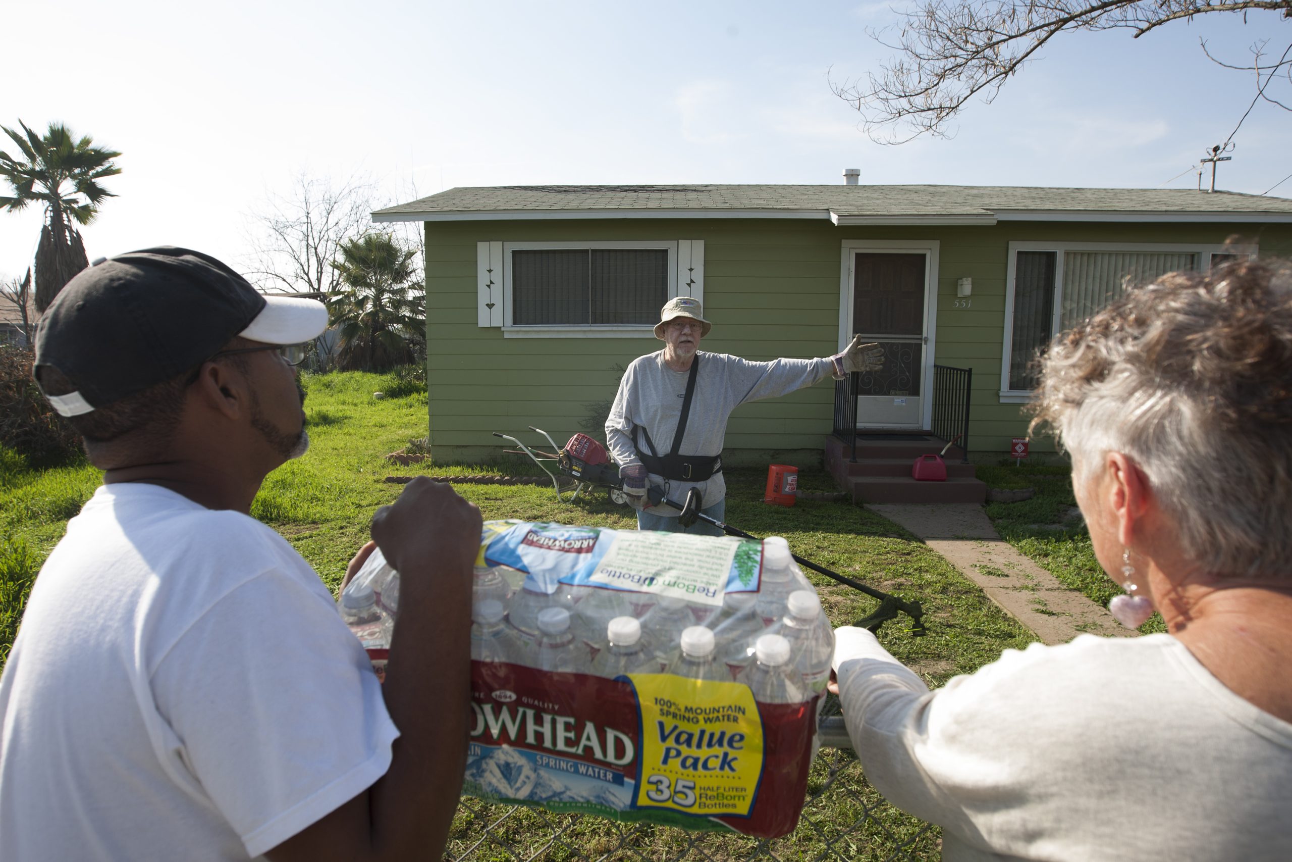 The three people identified in the caption are standing outside a small, green ranch-style house, two holding a pack of water bottles.