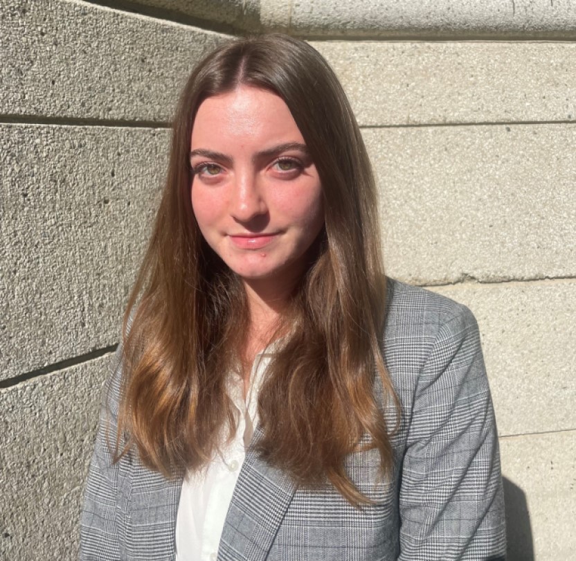 Katherine Hapgood has long brown hair and is wearing a black and white plaid jacket and a white blouse.