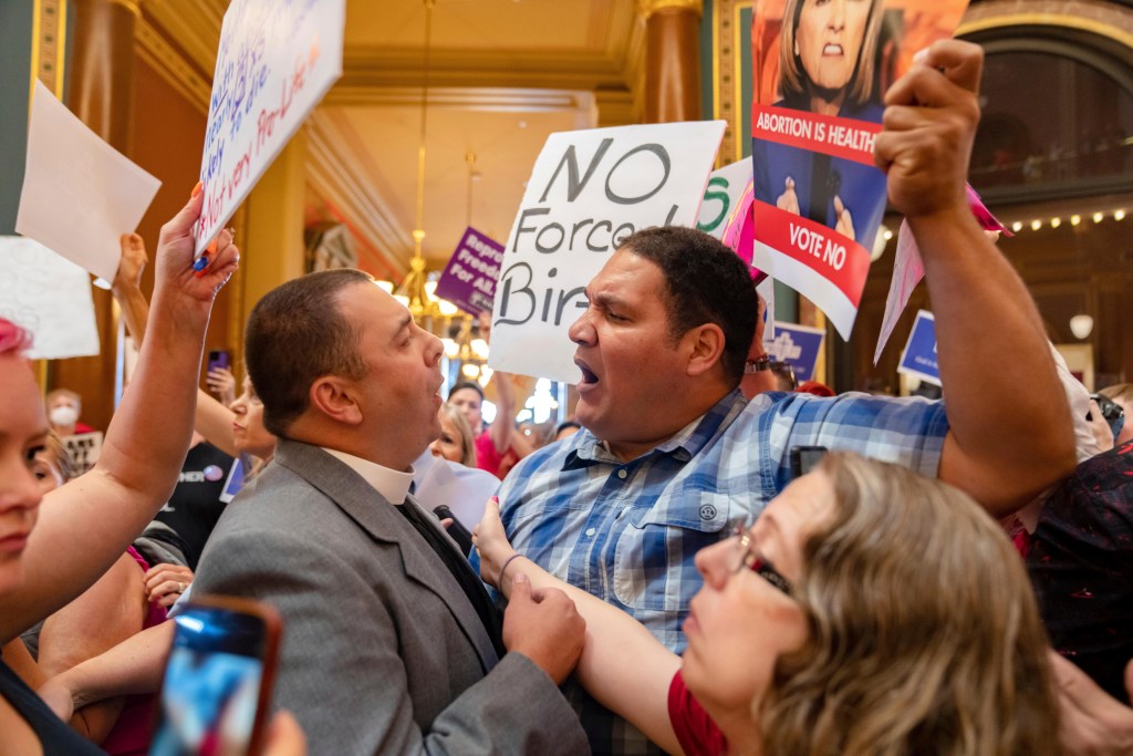 Michael Shover has a heated discussion with Ryan Maher, who is holding a protest sign, while a woman a woman puts her hand between the two men to try to break up the argument. They are surrounded by numerous protesters holding up signs.