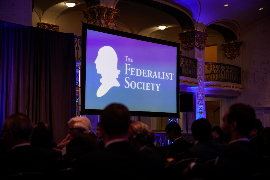 A huge monitors has the words "The Federalist Society" on it, while spectators sit in chairs mingling.