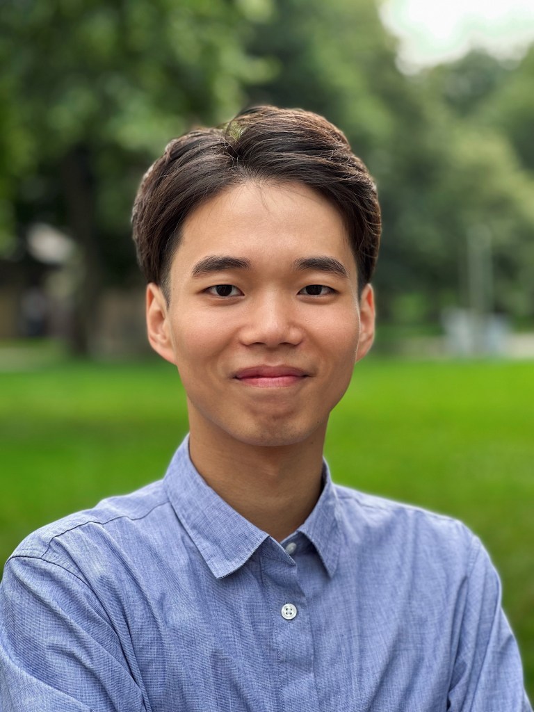 Jovi Dai has short brown hair and is wearing a blue shirt. He stands in front of a green lawn and trees.