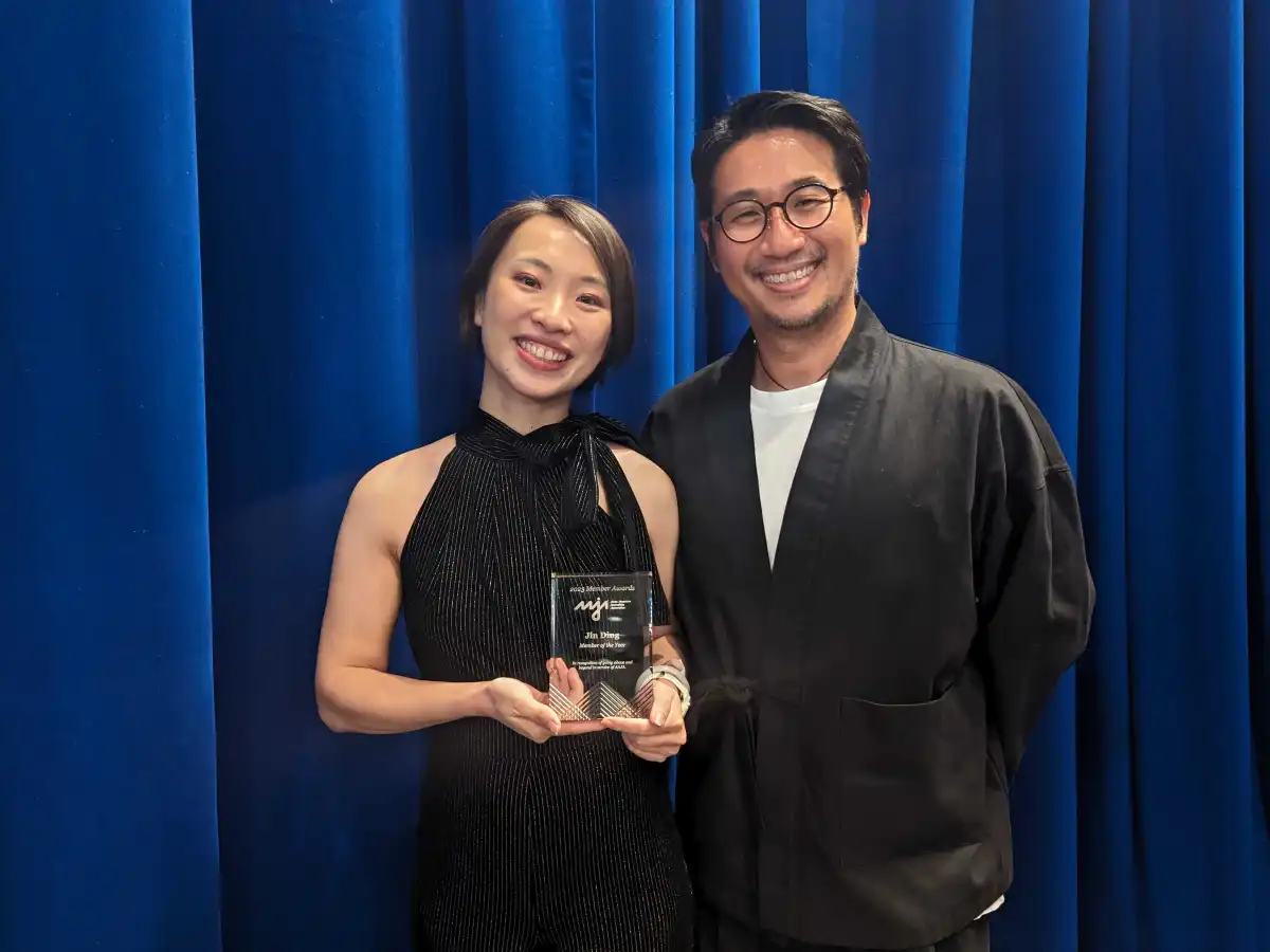 A woman in a black dress standing in front of a blue curtain and holding a glass plaque stands next to a man in a black sport coat and T-shirt.