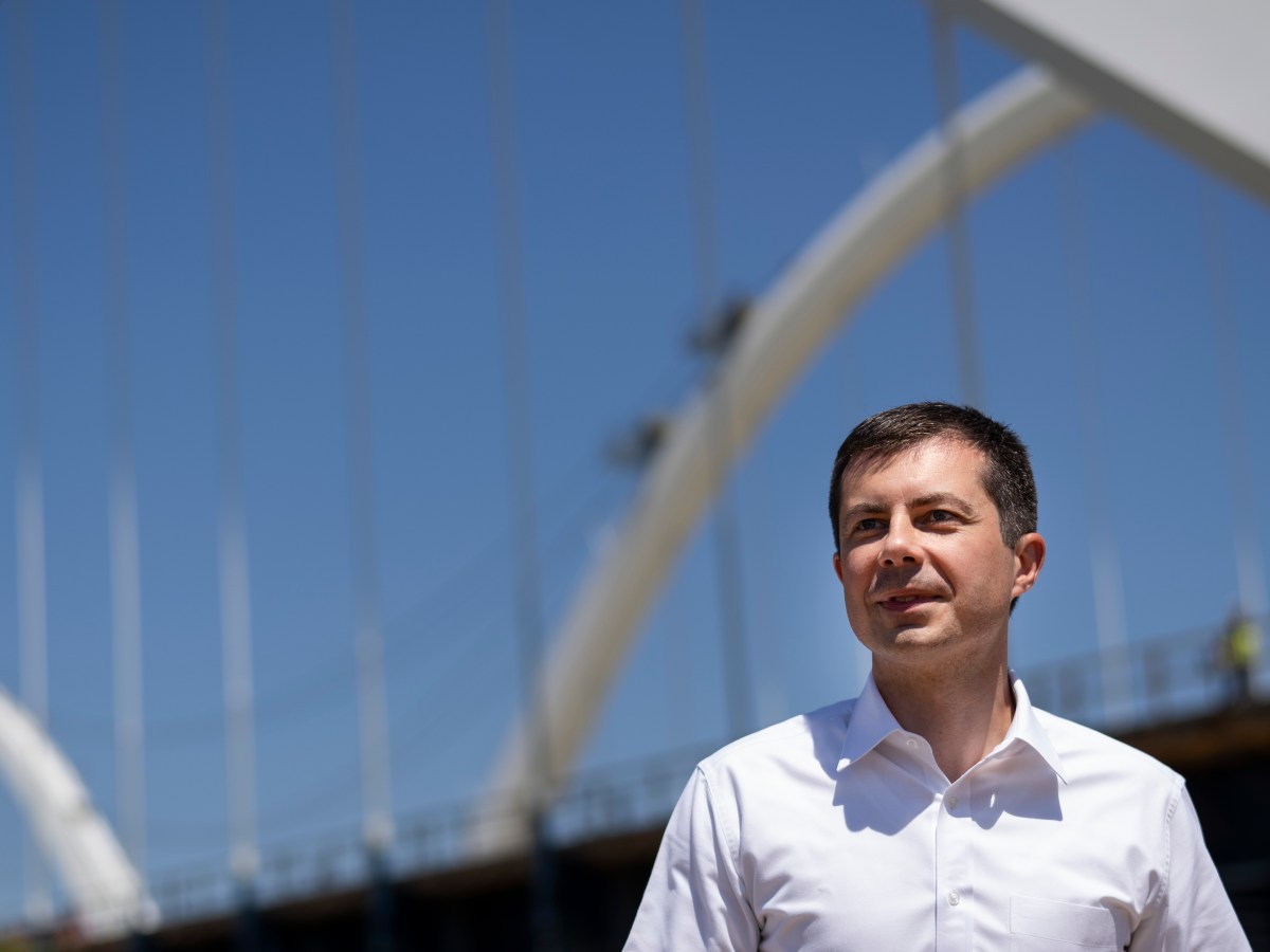Pete Buttigieg stands in the foreground wearing a white buttoned-down shirt. In the background is the Frederick Douglass Memorial Bridge, which is in the urban area of Washington D.C.