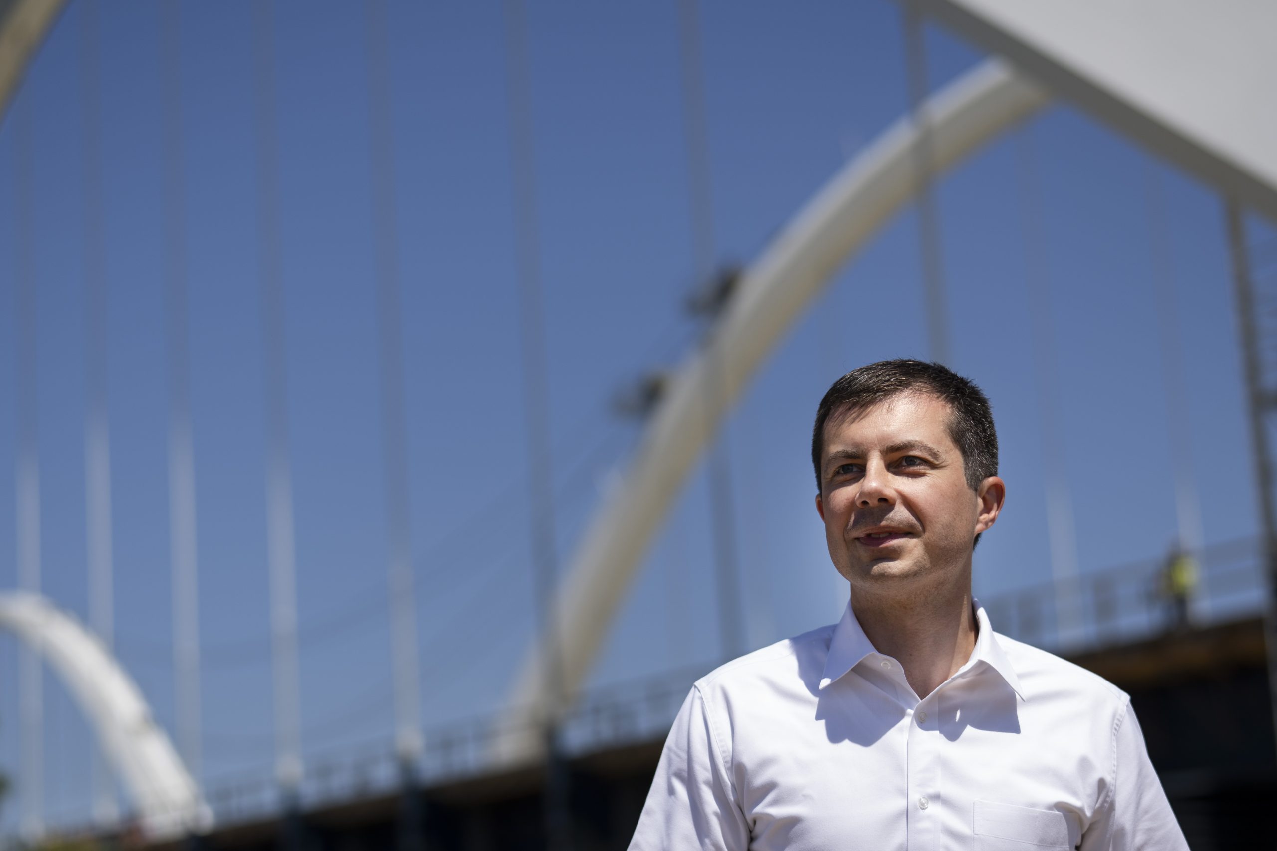 Pete Buttigieg stands in the foreground wearing a white buttoned-down shirt. In the background is the Frederick Douglass Memorial Bridge, which is in the urban area of Washington D.C.