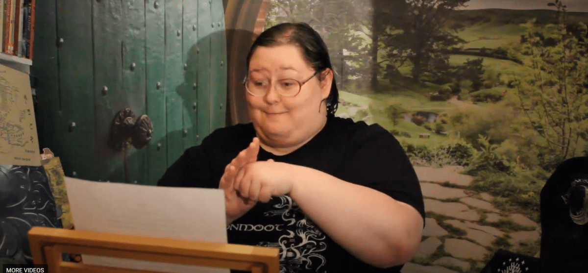 Hailey Simon uses American Sign Language to tell her story about what home means to her. She has short dark hair and wears glasses. She is reading from a paper on a wooden stand. Behind her is a picture of a stone path and trees. To her right is a green door with a knob in the middle.
