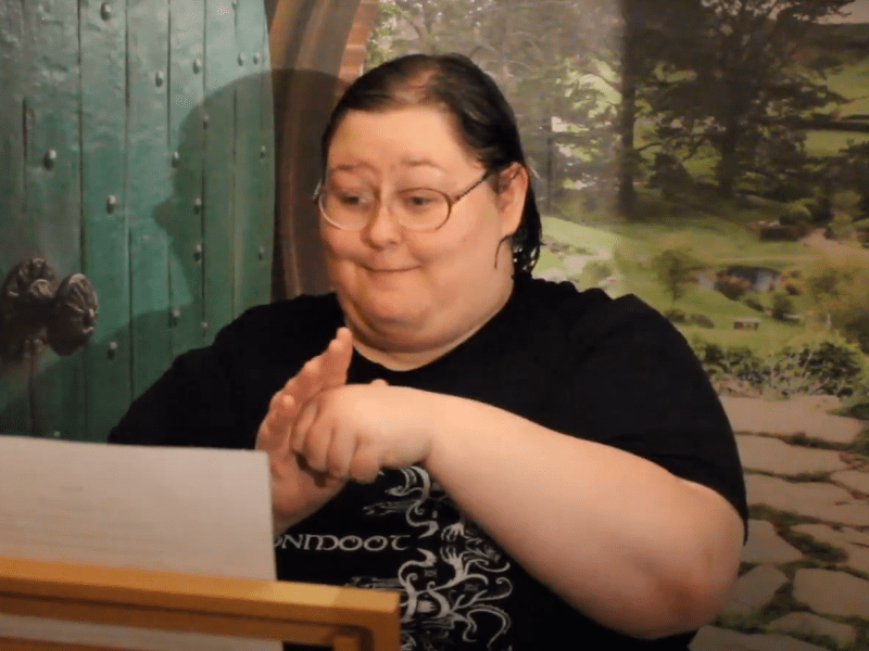Hailey Simon uses American Sign Language to tell her story about what home means to her. She has short dark hair and wears glasses. She is reading from a paper on a wooden stand. Behind her is a picture of a stone path and trees. To her right is a green door with a knob in the middle.