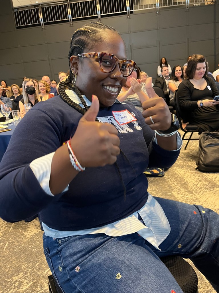 A smiling, seated woman in a blue sweater and glasses and a name tag that says, "Ashley Clarke," gives two thumbs up to the camera.