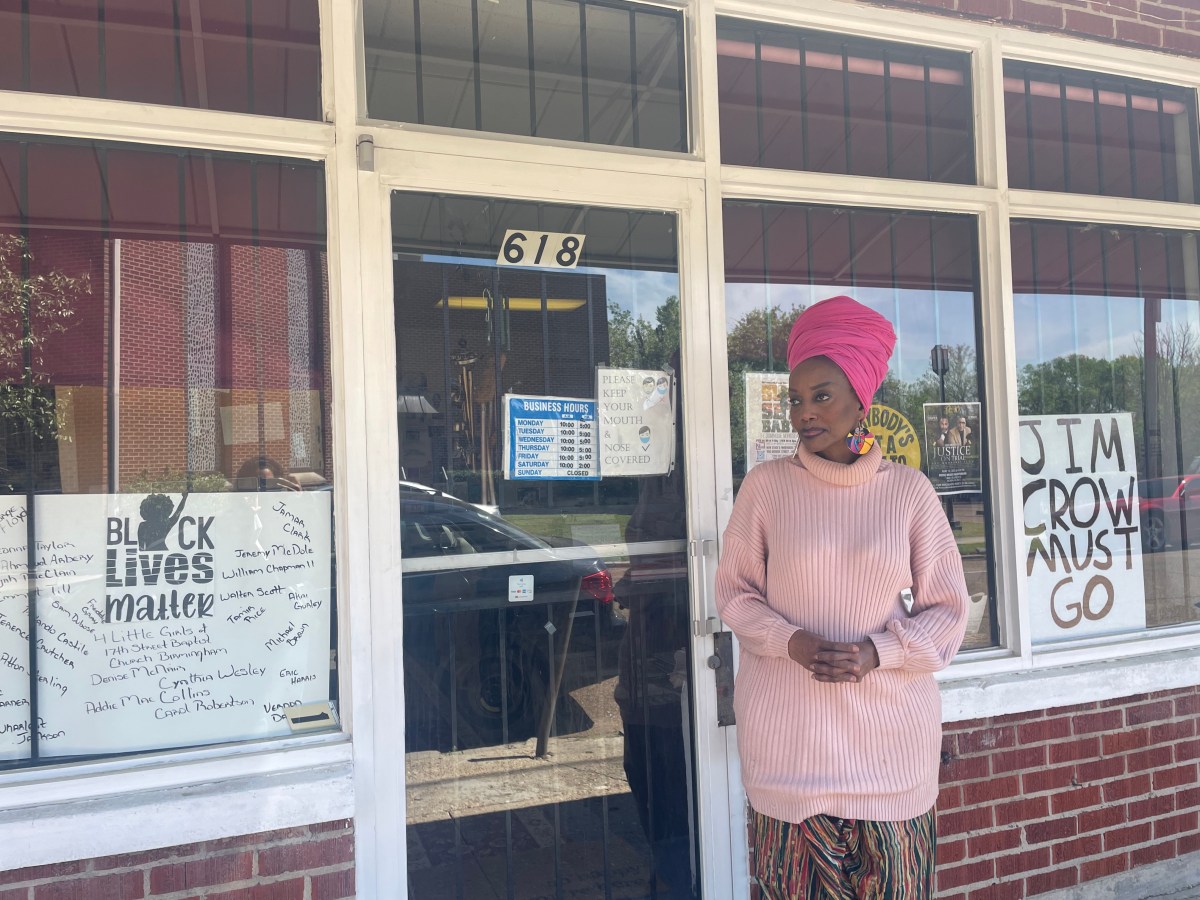 Maati Jone Primm stands in front of her store. She is wearing a pink outfit, and she has two signs in the windows of her store. One says "Jim Crow Must Go" and the other says "Black Lives Matter."