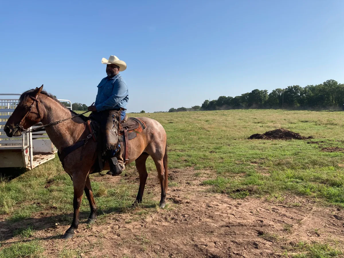 Nate dressed in a cowboy hat, blue hat and pants sits on a horse. His large farm with tall trees is in the background.