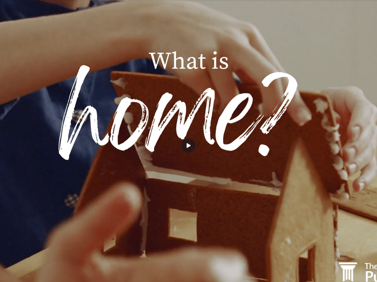 A screenshot from a video showing a person about to put the roof on a gingerbread house. The person is wearing a blue shirt.