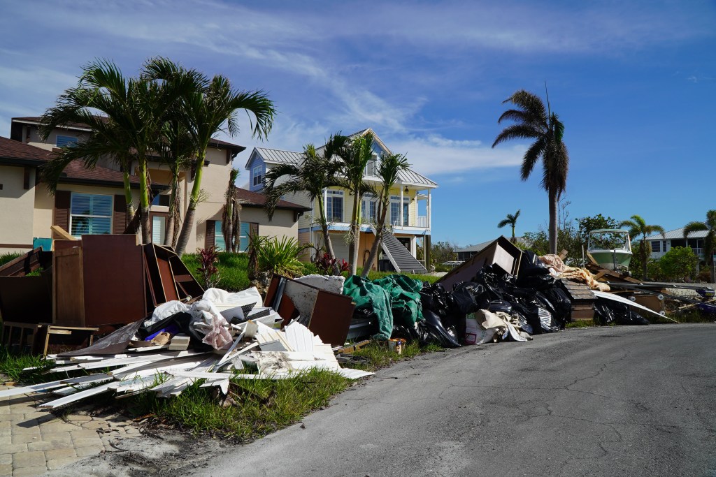 Piles of debris, including furniture, sit on the curb waiting to be picked up. A yellow building stands behind the debris–  it was one of a few structures left standing in Fort Myers Beach after Hurricane Ian.
