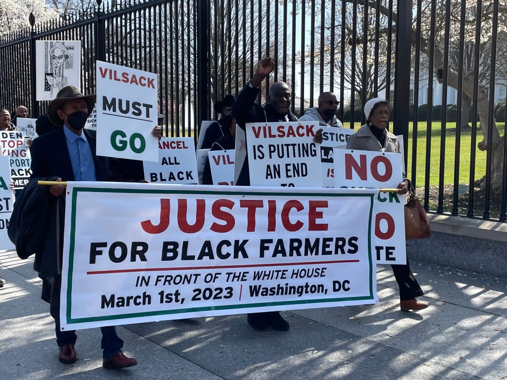Several protesters walk in front of the White House gate with signs that read "Justice for Black farmers. In front of the White House March 1st, 2023. Washington, DC."