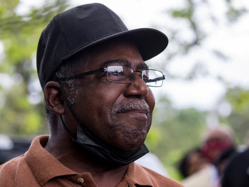 Eddie Slaughter tears up. He is wearing a black baseball cap, mask around his chin and eye glasses.