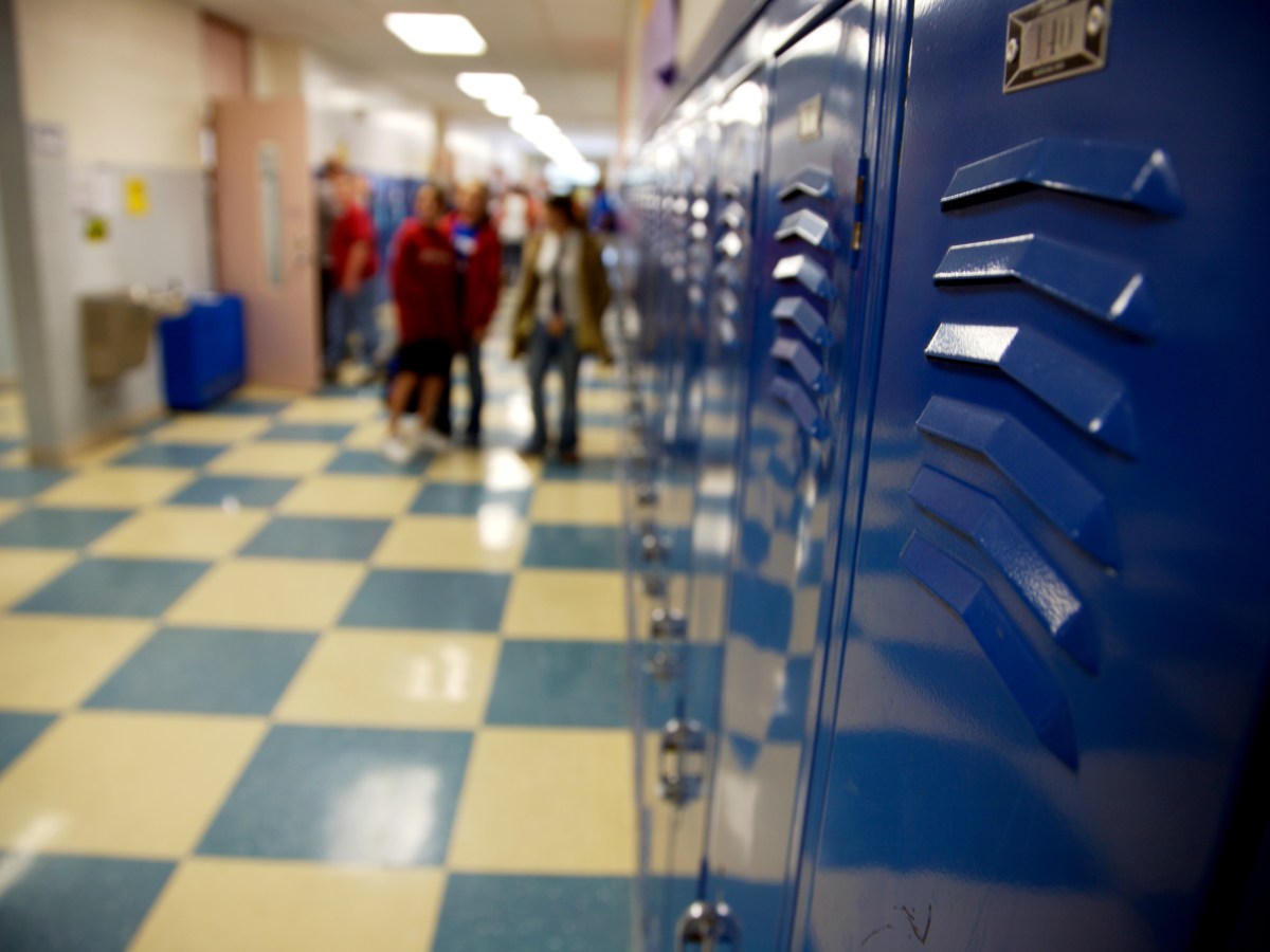 Blue school lockers are in focus in the foreground of the image showing a school hallway. In the background, out of focus, several people stand near each other and one person walks into a classroom.