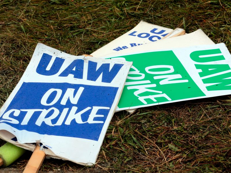 Three UAW On Strike picket signs stacked on top of each other on the grass. The topmost sign has blue text, and the one underneath it is in green. Both say "UAW ON STRIKE."