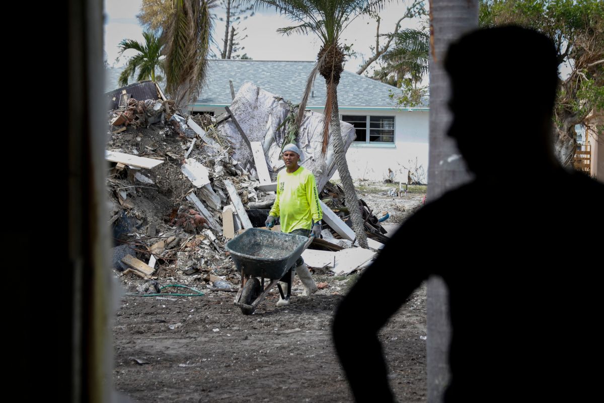 A worker with a wheelbarrow walk along a hurricane cleanup site. There is shadow of another person in the foreground.