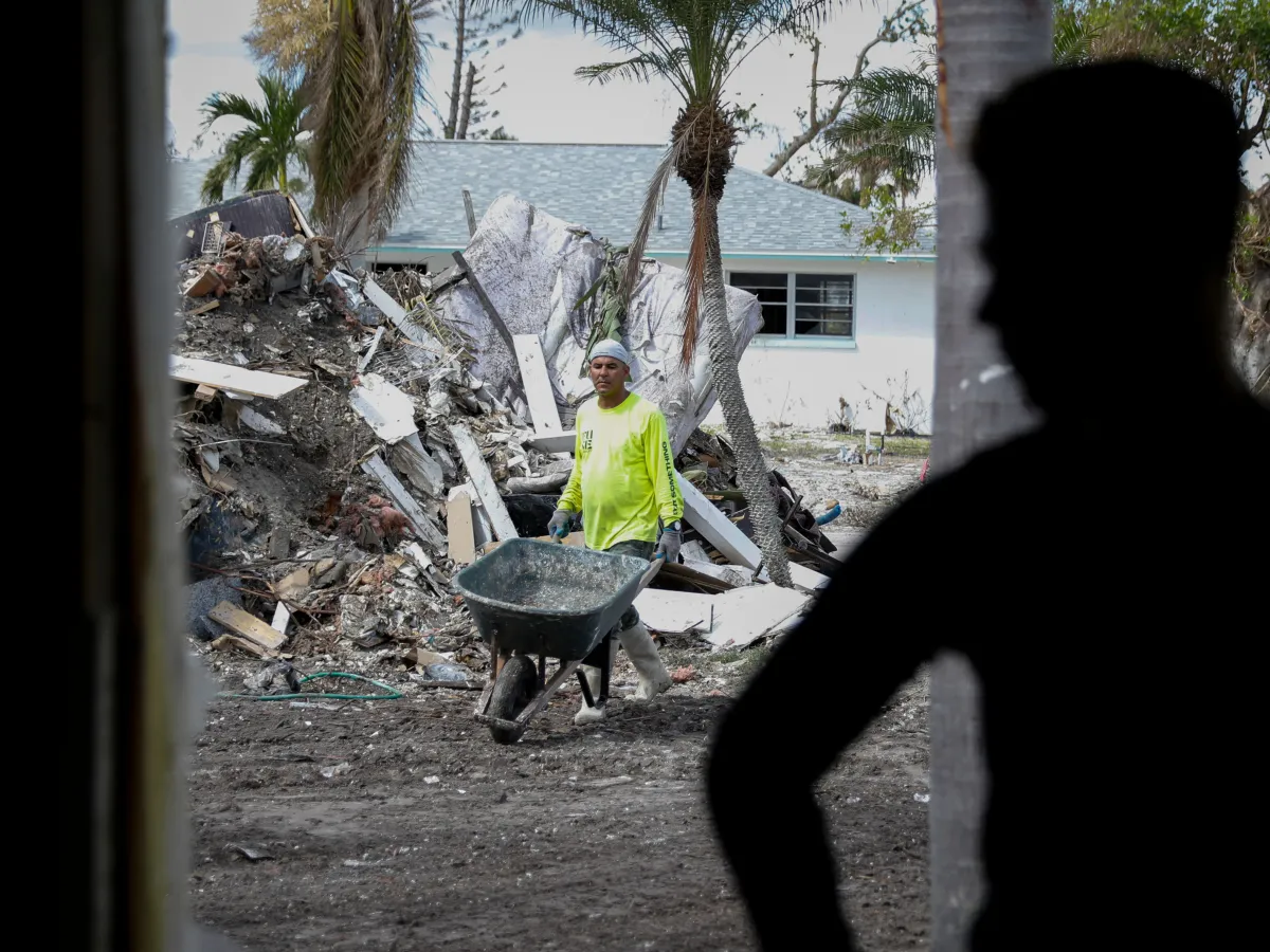 A worker with a wheelbarrow walk along a hurricane cleanup site. There is shadow of another person in the foreground.