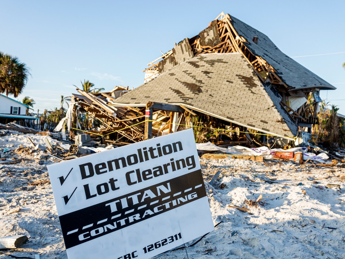 A demolition service sign is planted in the sand in front of a damaged building.