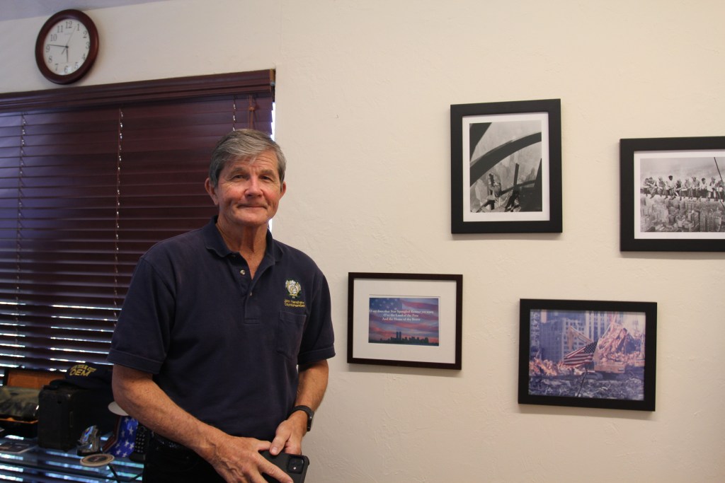 John Henshaw stands next to a photo gallery of 9/11 photos on the wall. 