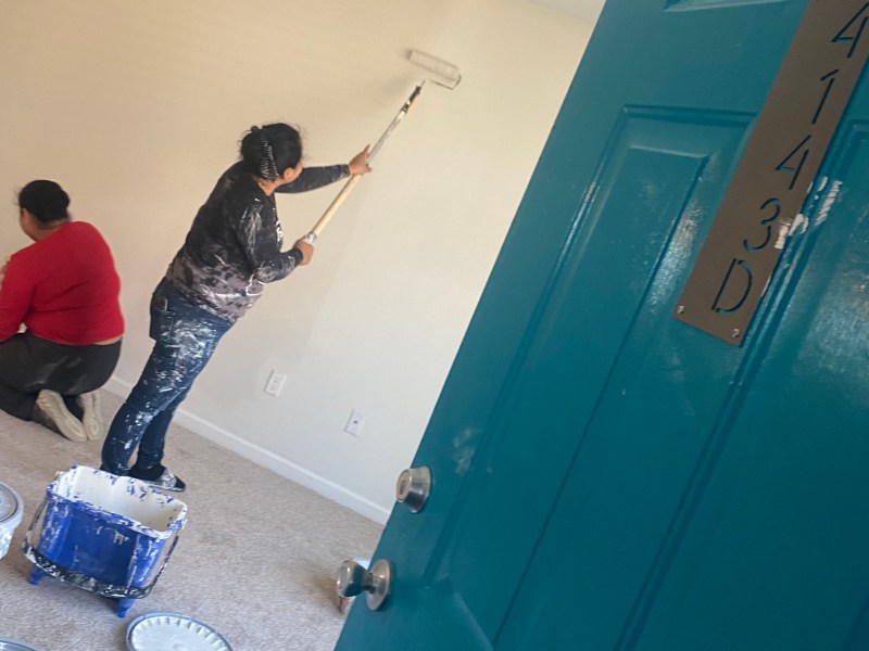 Disaster restoration worker Jenny, dressed in a black shirt and jeans, and her sister Abigail, dressed in an orange shirt and black pants, can be seen through a teal doorway, painting a vacant apartment at the Mayfair Apartment Homes in New Orleans with rollers and brushes, facing away from the camera. The right side of Jenny’s body is covered in splotches of white paint.