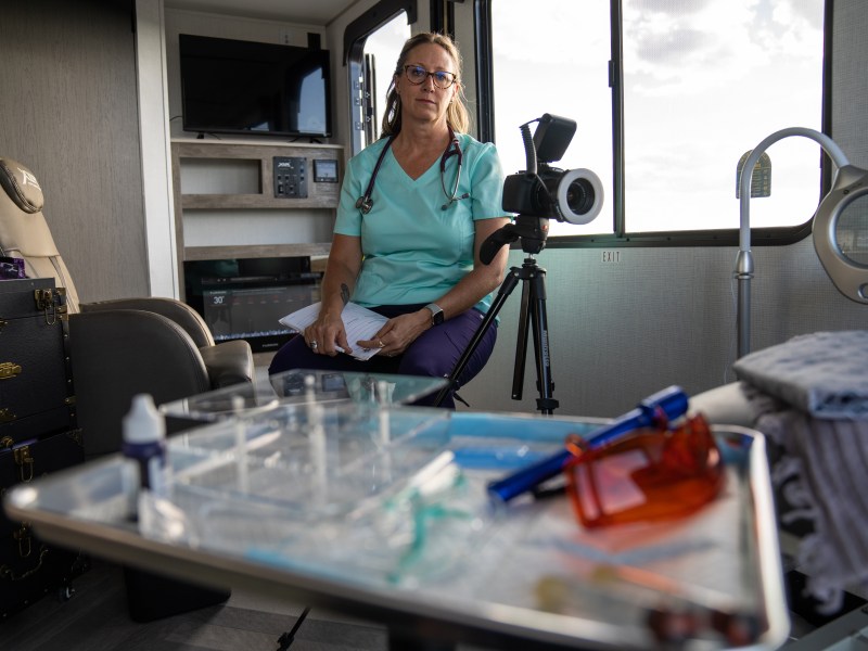 Norah Lusk is dressed in blue medical scrubs with a stethescope draped around her nck. She is sitting in a mobile medical unit.