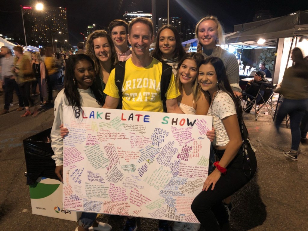 Ron Blake is surrounded by several people as he holds a sign that says "Blake Late Show".