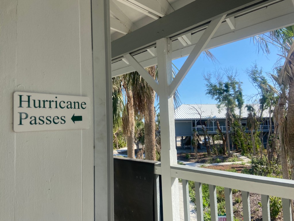 A white sign with black letters that reads “Hurricane passes” hangs outside Sanibel City Hall.  The damage from Hurricane Ian’s wind can be seen in the battered trees in the background. Sanibel officials limited access to the wealthy community by issuing hurricane passes to workers involved in the recovery effort.  
