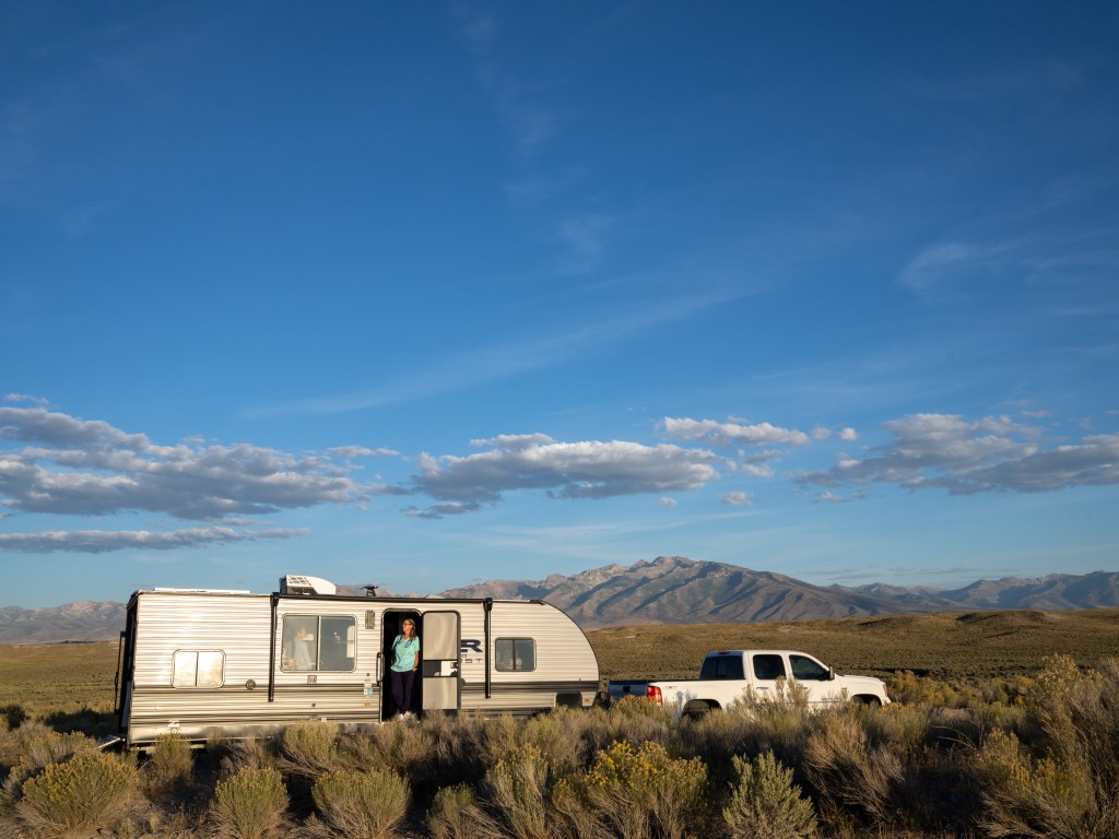 With mountains in the background, a pickup truck sits stationary with a trailer attached to it. Standing in the doorway of the trailer is Nora Lusk with her blue scrubs.