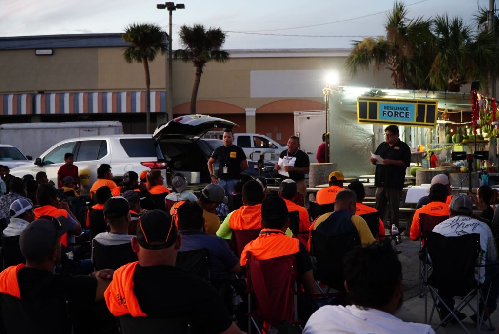 Resilience Force activist and former disaster restoration worker, Mariano, stands in the middle of his two co-workers holding documents while speaking to workers in November 2022, two months after Hurricane Ian devastated the region. Scores of disaster restoration workers, some dressed in bright orange long sleeve shirts, listen during the first worker meeting organized by Resilience Force in Fort Myers, post-Ian.