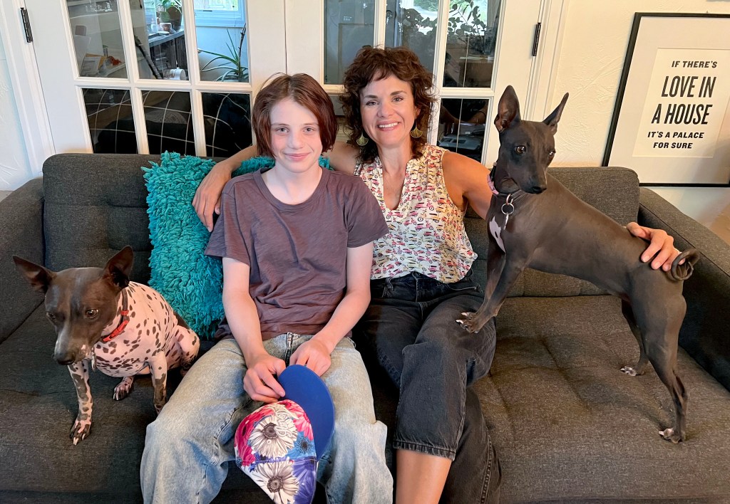 Maddie Maes and her mother, Kristin Vera, pose for a photo on their couch at home in Fort Collins. Maddie is smiling and wears a brown t-shirt, faded jeans and is holding a flowered ball cap in her hands. Her mother also is smiling and has her arm around Maddie. Her mother wears a patterned tank top and black jeans. Kristin also has her hand on a small brown dog on her left that looks like a small Doberman pincer. Next to Maddie is another dog that is pinkish with brown spots and has a brown head. Behind the couch are two french doors and on the wall is a framed picture that says: "If there's love in a house It's a palace for sure.