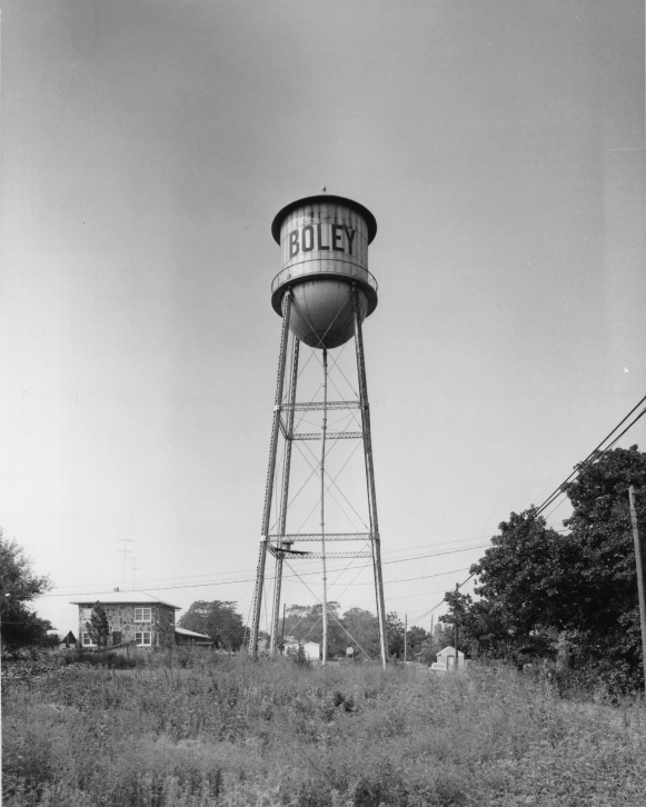 A black and white image of a water tower that has the word Boley on it. The water tower sits in an empty field. Behind it is a small building in the distanc.