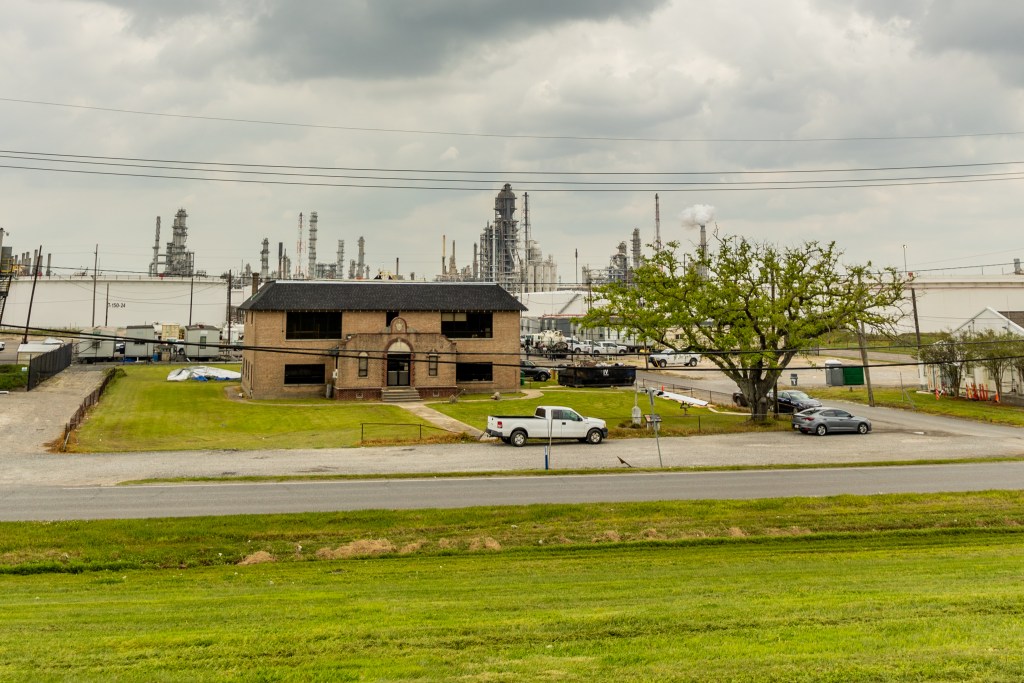 Gray clouds swirl over gray refinery towers. In the foreground, close to industrial infrastructure, is the former school, brown brick, with a tree nearby.