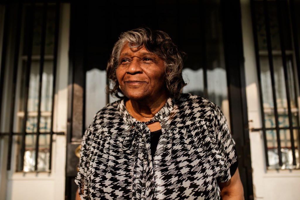 Erma Barnes smiles as she stands in front of her house, which has a black door, while wearing a black and white top.
