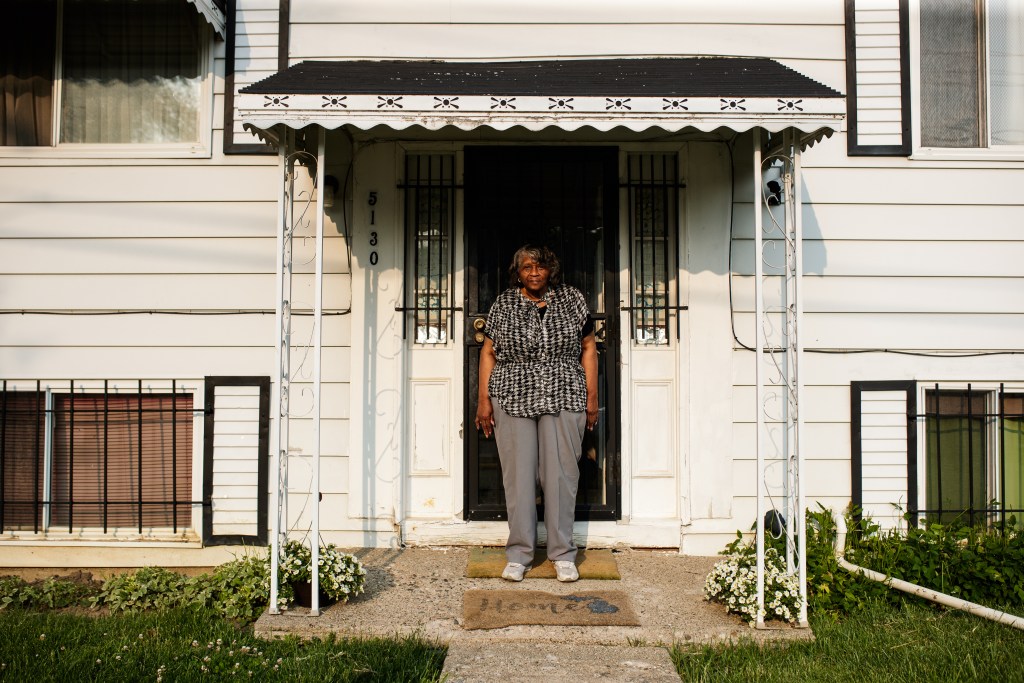 Erma Barnes is standing on her front porch. Her home has light-colored siding and a welcome mat that says "Home."