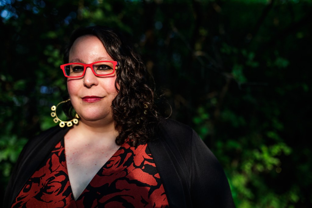 Mona Munroe-Younis smiles while wearing a black and red top, red glasses and gold hoop earrings.
