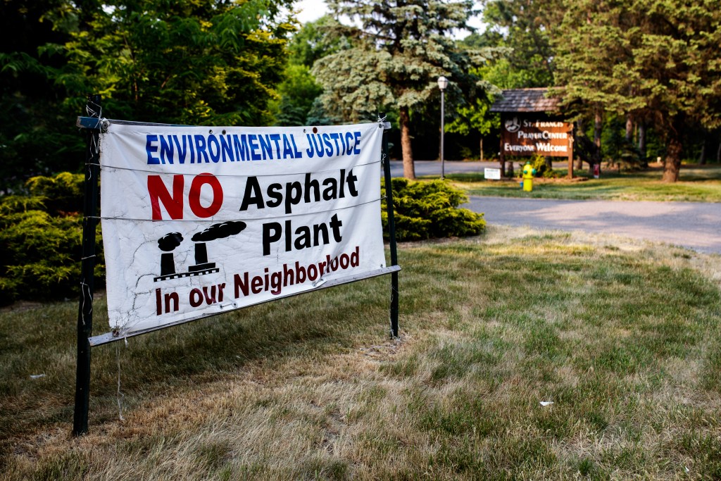 A sign, pitched in the grass, says Environmental Justice. No Asphalt Plant in our Neighborhood.