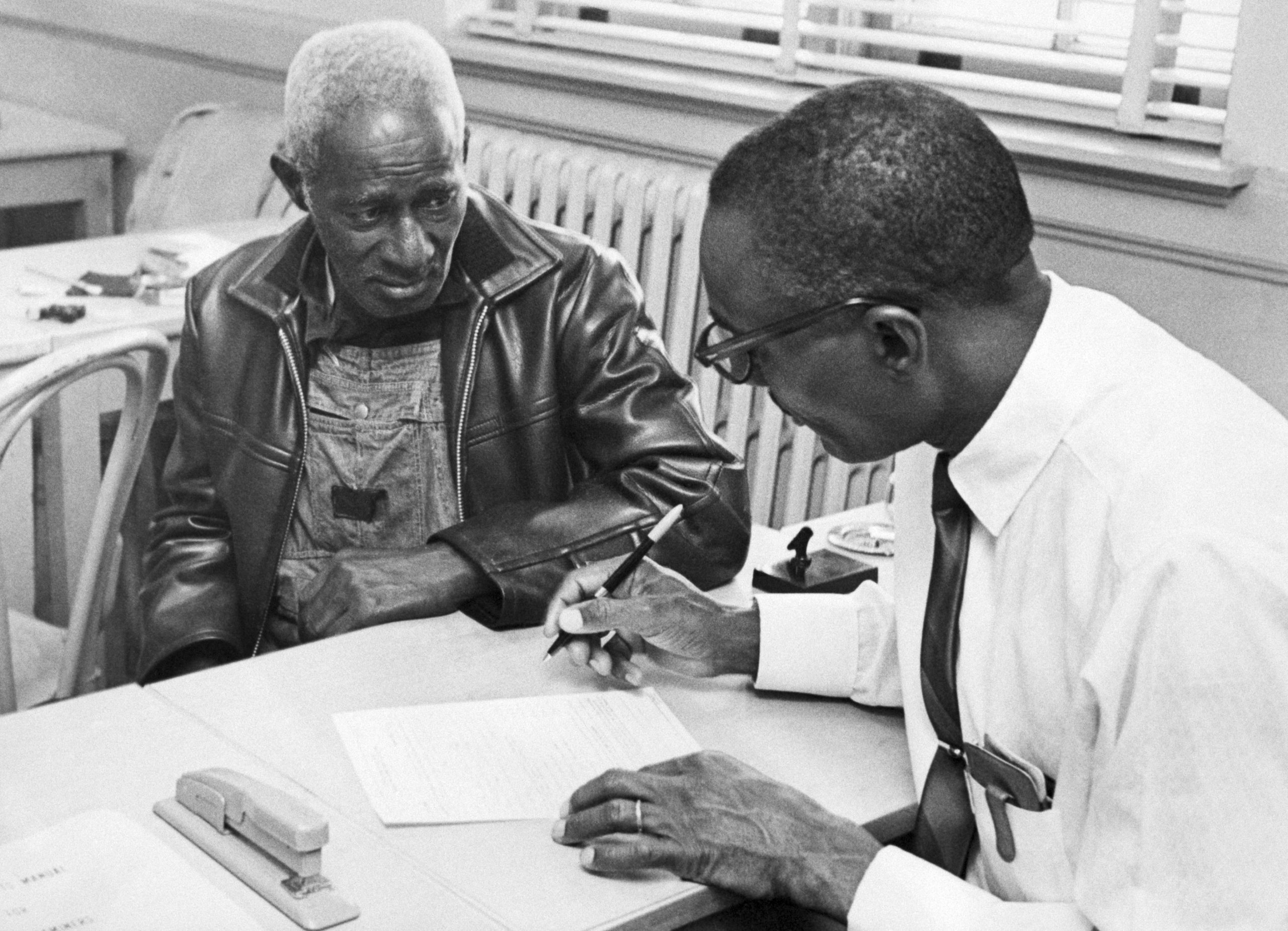 Two Black men sit at a table, going over paperwork together. There's a window in the background.