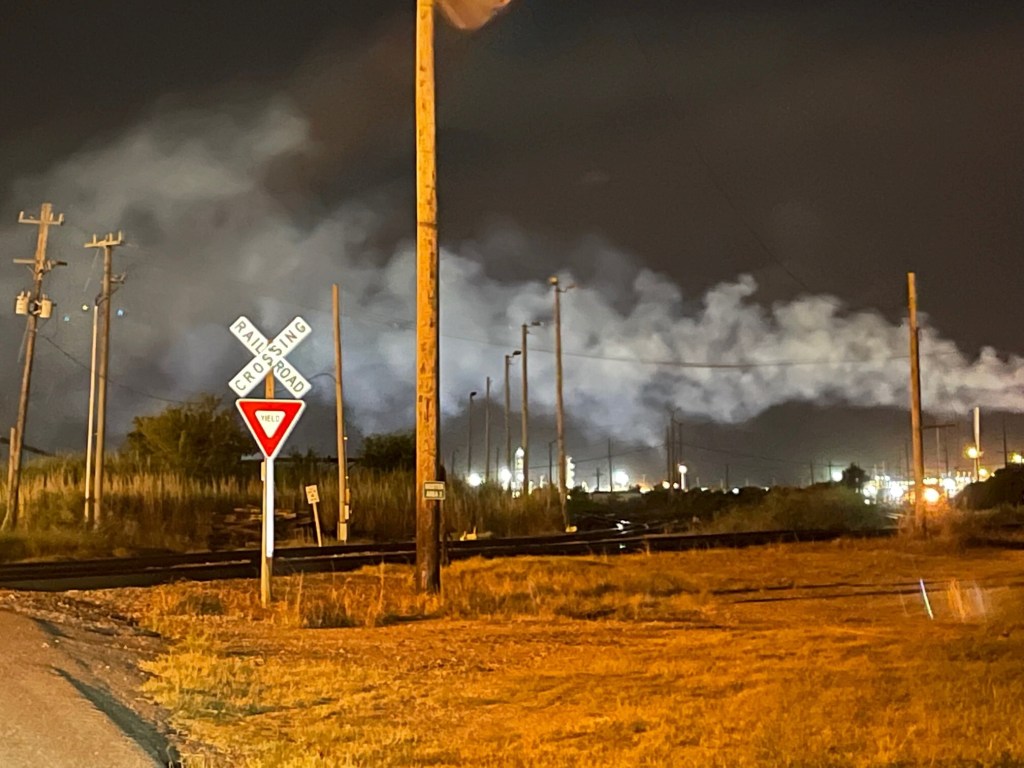 A plume of emissions that look like smoke rolls across a dark sky against the lights of the Oxbow Calcining plant. Train tracks and a railroad crossing sign are in the foreground.