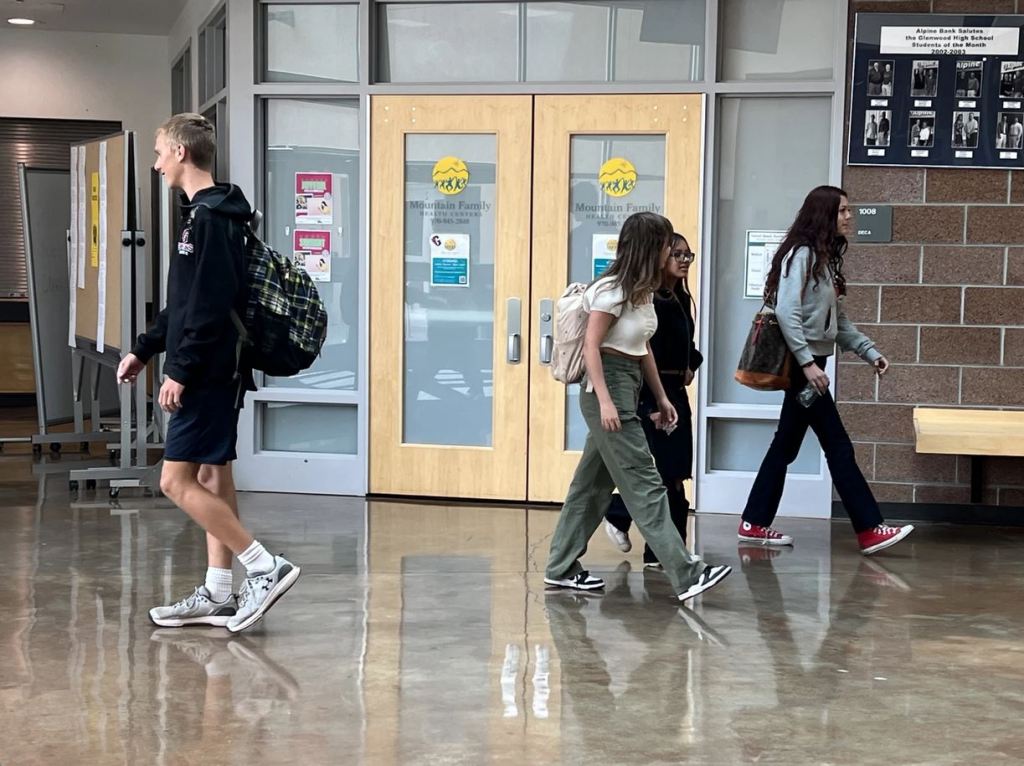 Students walking in school hallway in front of an office that says "Mountain Family" on the doors. A boy wearing a black hoodie and black shorts and a plaid backpack walks one way. Three girls walk the other way