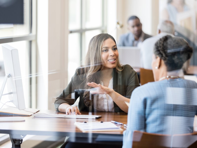 Two Black women discuss banking in this photo, taken in a corporate setting with light coming in through windows