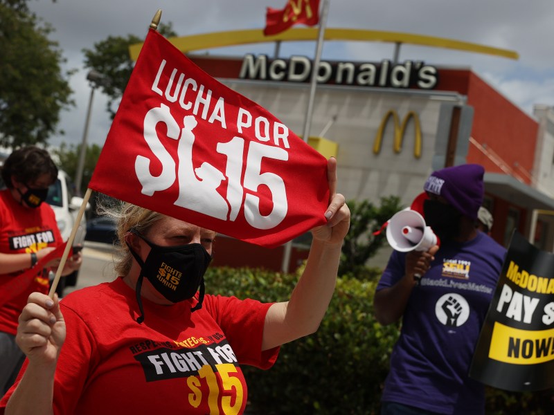 A woman in a red tshirt protests outside of a McDonalds. She holds up a red sign that reads "Lucha por $15"