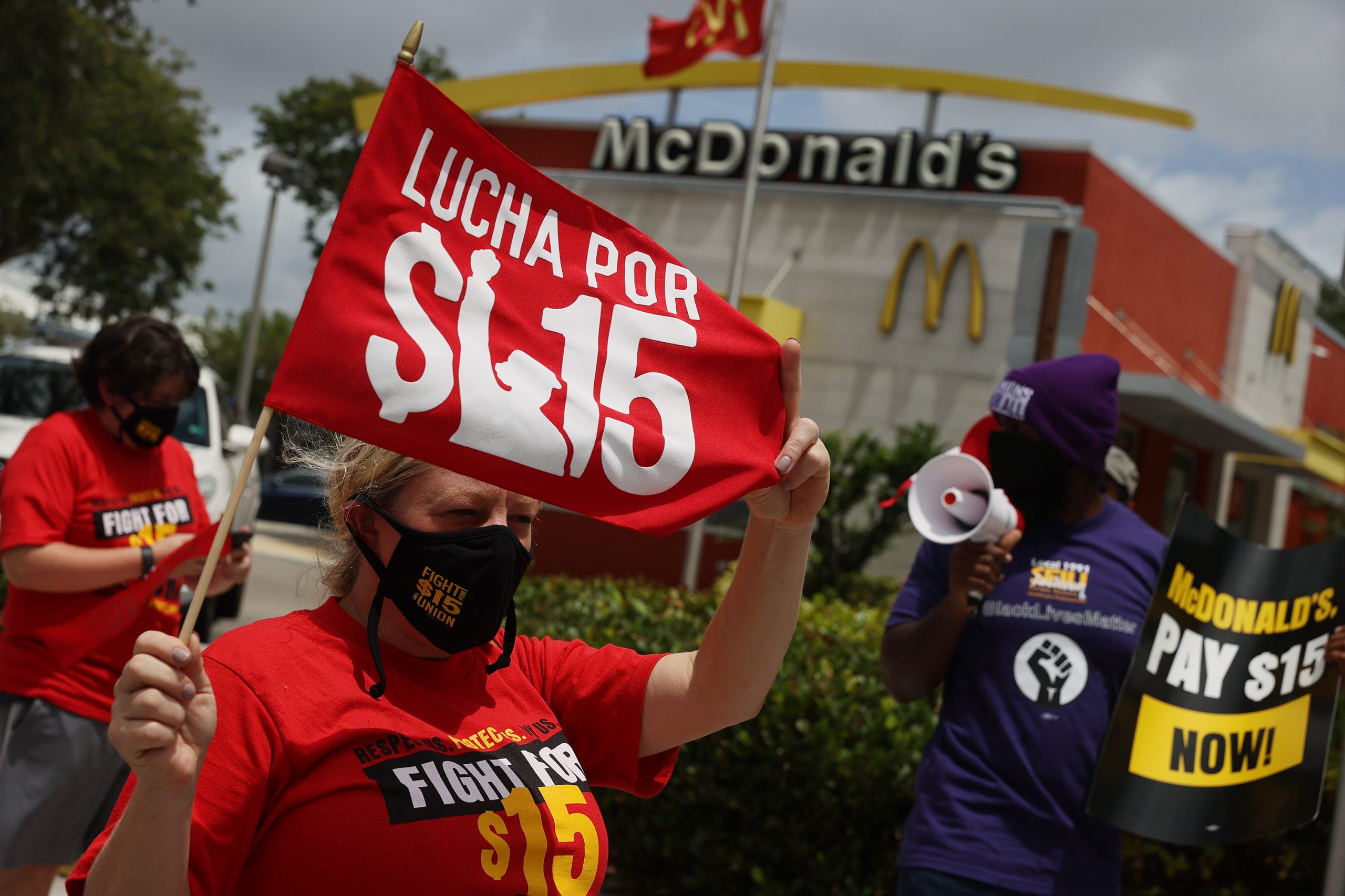 A woman in a red tshirt protests outside of a McDonalds. She holds up a red sign that reads "Lucha por $15"