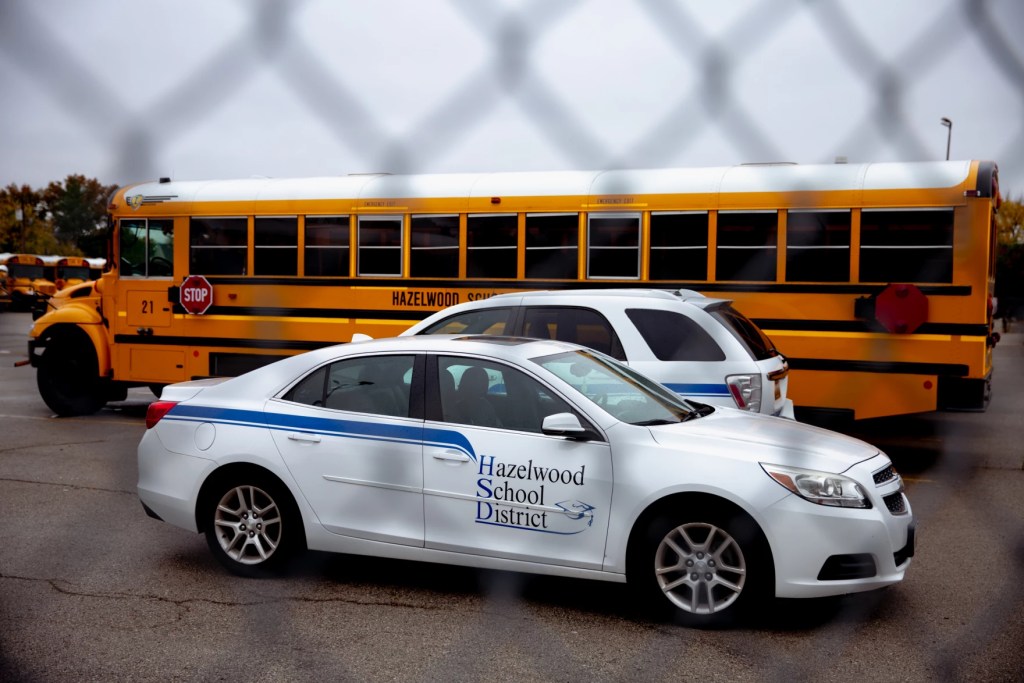 In this photo, yellow school buses are visible behind two cars with the school district's name on them, all viewed through chain-link fencing.