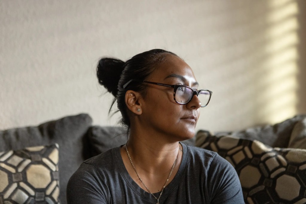 Elizabeth Ramirez, mother to three children, sits at home. She looks to her left. She wears a gray top, a necklace and brown tortoise shell glasses. Her dark hair is pulled up.