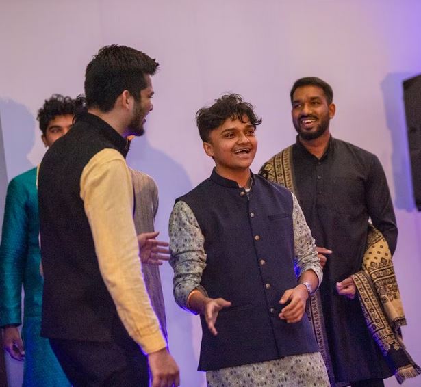 Students at Georgia State University talk at a Diwali fashion show. Four students are talking. In the center is a young man wearing a blue fest and is smiling. Behind him other students are smiling.