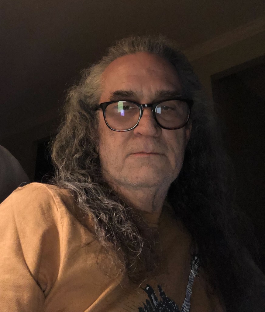 Jacques LeBourgeois is looking directly into the camera with a serious expression. He has long gray hair, is clean-shaven and is wearing glasses.