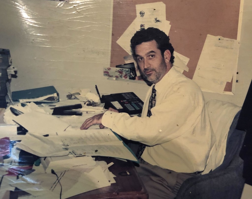 Jacques LeBourgeois is sitting at a paperwork-covered desk, a corkboard with paperwork on it behind him. He's turned to glance at the camera and has short, dark hair, a beard and mustache.