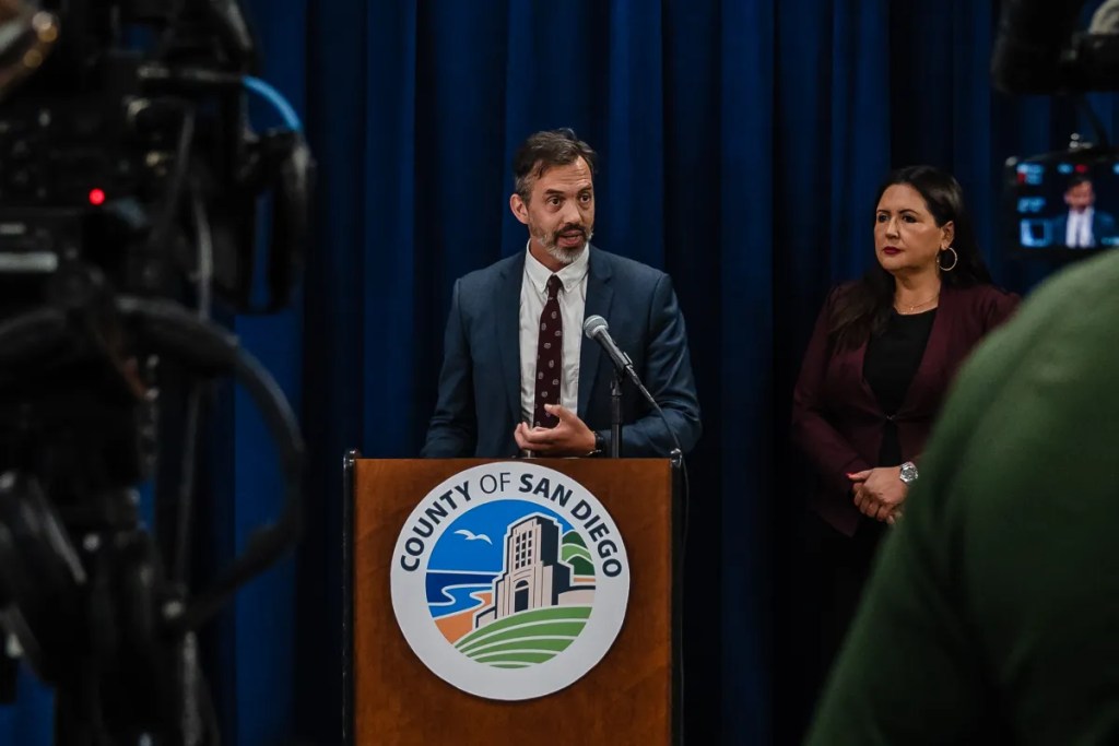 County Behavioral Health Services Director Luke Bergmann speaks to members of the media about the CARE Act program at the County Administration Center. He stands at a wooden podium with a San Diego County logo on it. He has dark hair and a beard. He wears a blue suit and a white shirt with a dark tie.